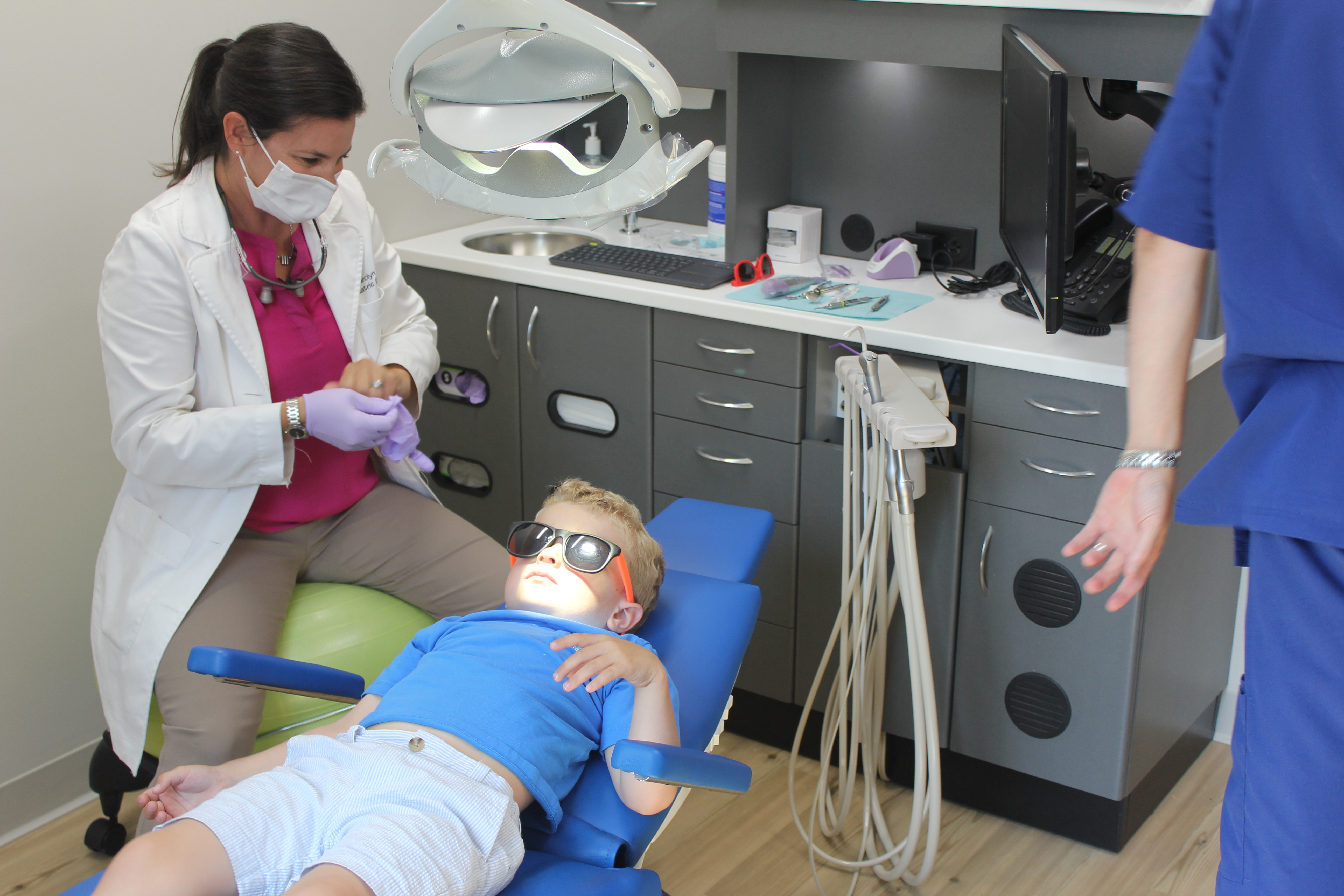 Child in sunglasses reclining in dental chair while pediatric dentist prepares tools in modern dental office, highlighting Coppe + Sears pediatric dentistry services in Lexington, MA.