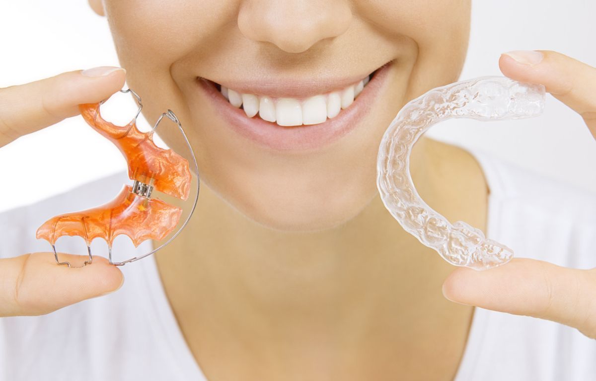 Smiling person holding orthodontic appliances, including a colorful dental retainer and a clear Invisalign aligner, representing options for orthodontic treatment at Coppe & Sears Orthodontics in Lexington, MA.