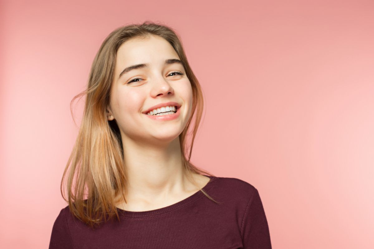 Smiling young woman with straight teeth and light brown hair against a pink background, representing the positive outcomes of orthodontic treatment at Coppe & Sears Orthodontics in Lexington, MA.