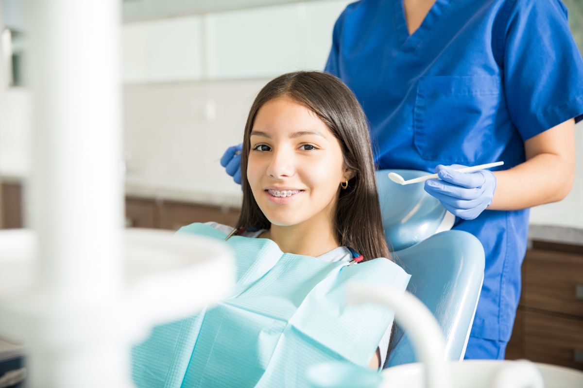 Smiling girl in dental chair with braces, pediatric dentist preparing tools, emphasizing orthodontic care at Coppe & Sears Pediatric Dentistry.