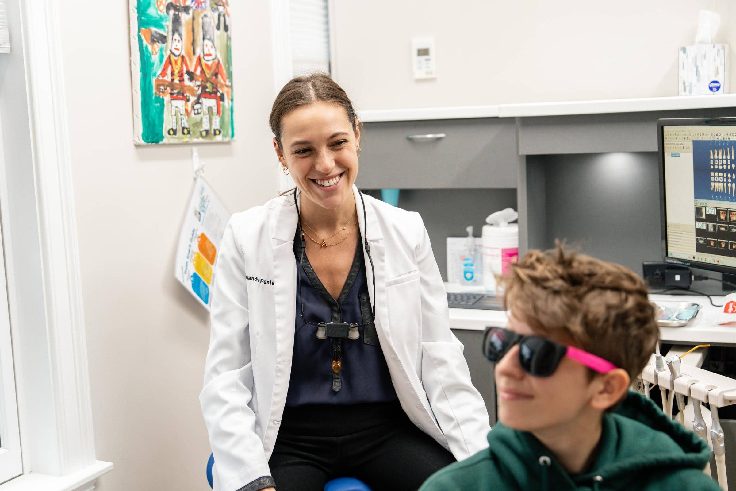 Smiling pediatric dentist in white coat interacting with a young patient wearing sunglasses in a dental office setting, reflecting the friendly environment of Coppe + Sears pediatric dentistry in Lexington, MA.