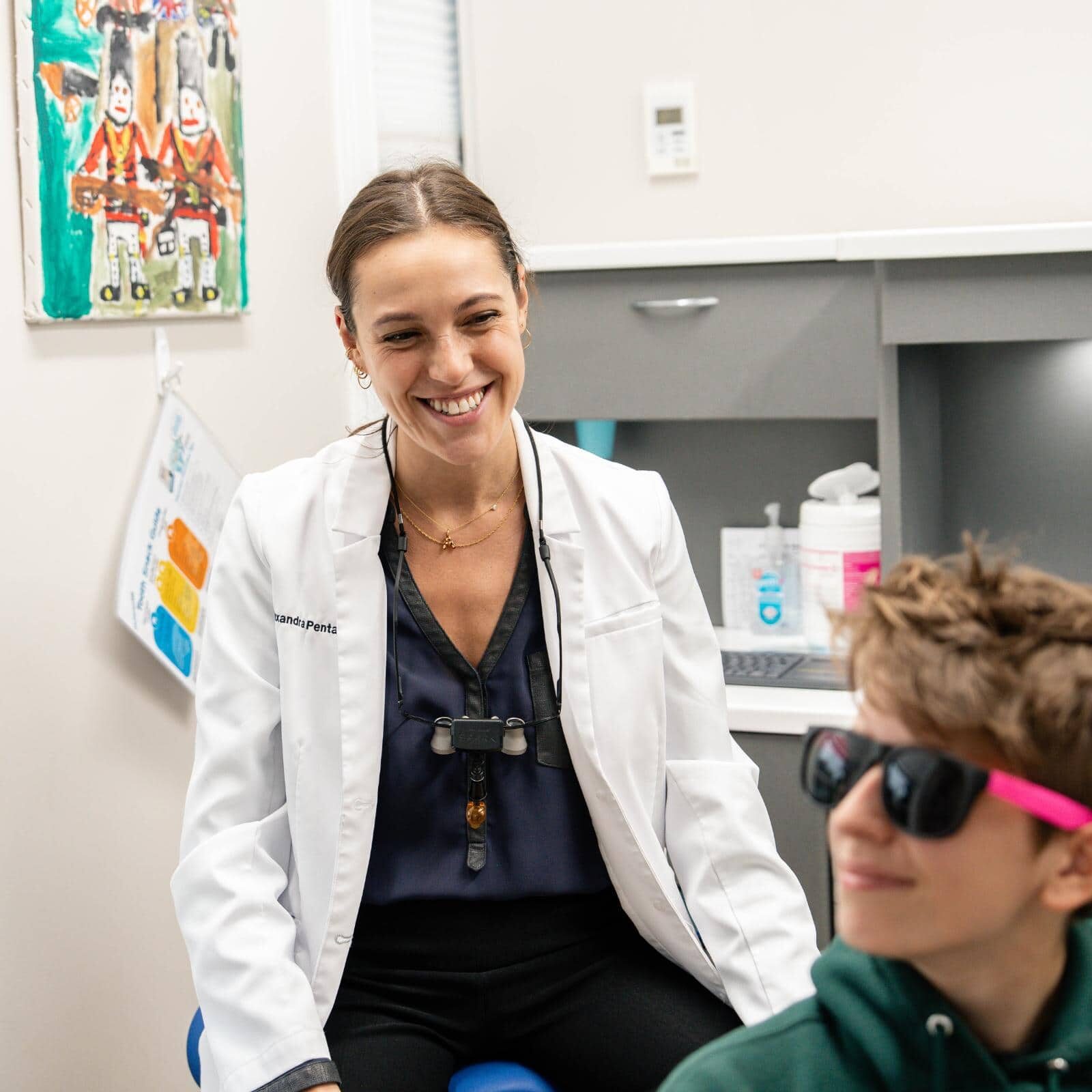 Smiling pediatric dentist in white coat engaging with young patient wearing sunglasses in modern pediatric dentistry office at Coppe + Sears in Lexington, MA.